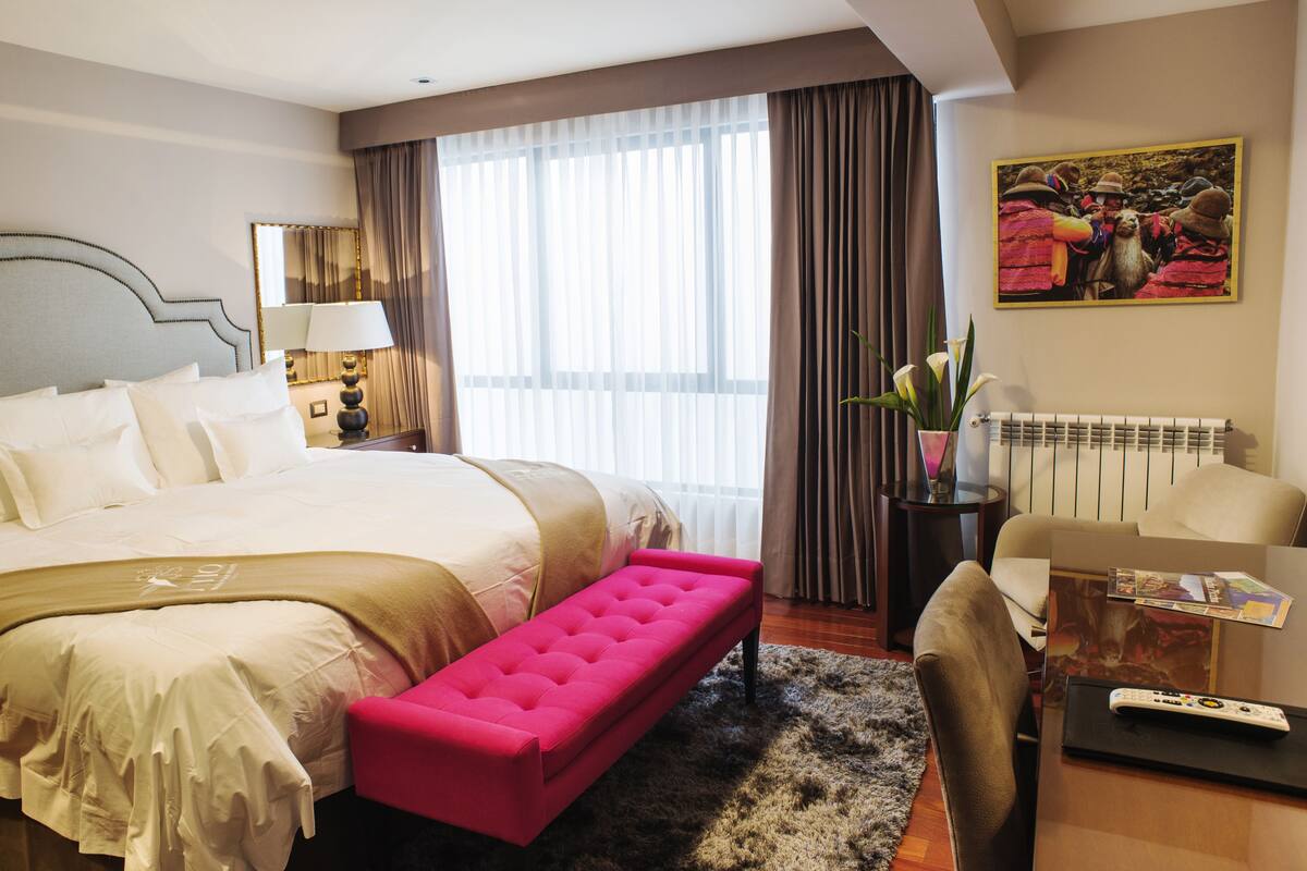 A hotel room with a large bed, a pink bench, a desk with a remote control, a chair, and a painting on the wall.