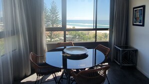 A room with a large window overlooking a beach, a round table with chairs, and a framed picture on the wall. - Princess Palm on the Beach (Palm Beach)
