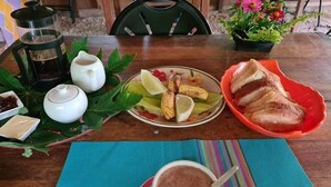 Una mesa con una cafetera, un plato de fruta cortada, un bol de pan y una taza de bebida caliente.