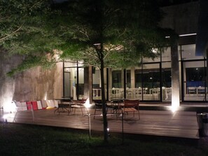 An outdoor seating area with tables and chairs, a tree, and a building with large windows in the background.