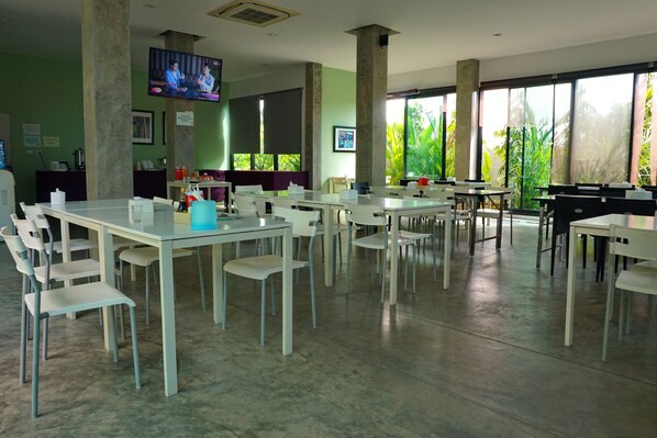 Un comedor espacioso con varias mesas y sillas, ventanas amplias con vistas a zonas verdes y un televisor fijado a un pilar.