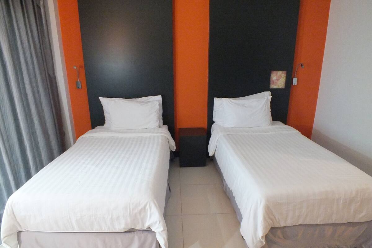 Ein Hotelzimmer mit zwei Einzelbetten, einem dunkel gefärbten Kopfteil und einem Vorhang auf der linken Seite.