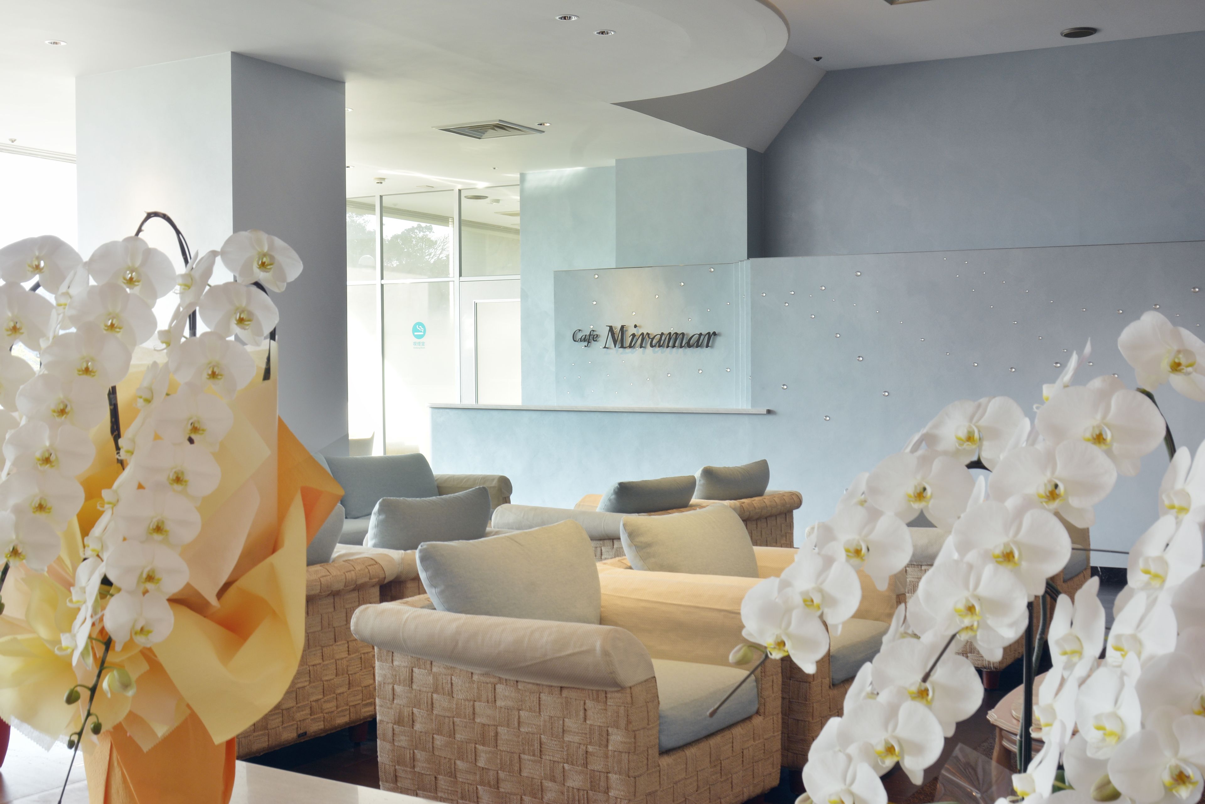 A modern lounge area with wicker sofas, white orchids, and a 'Cafe Miramar' sign on the wall.