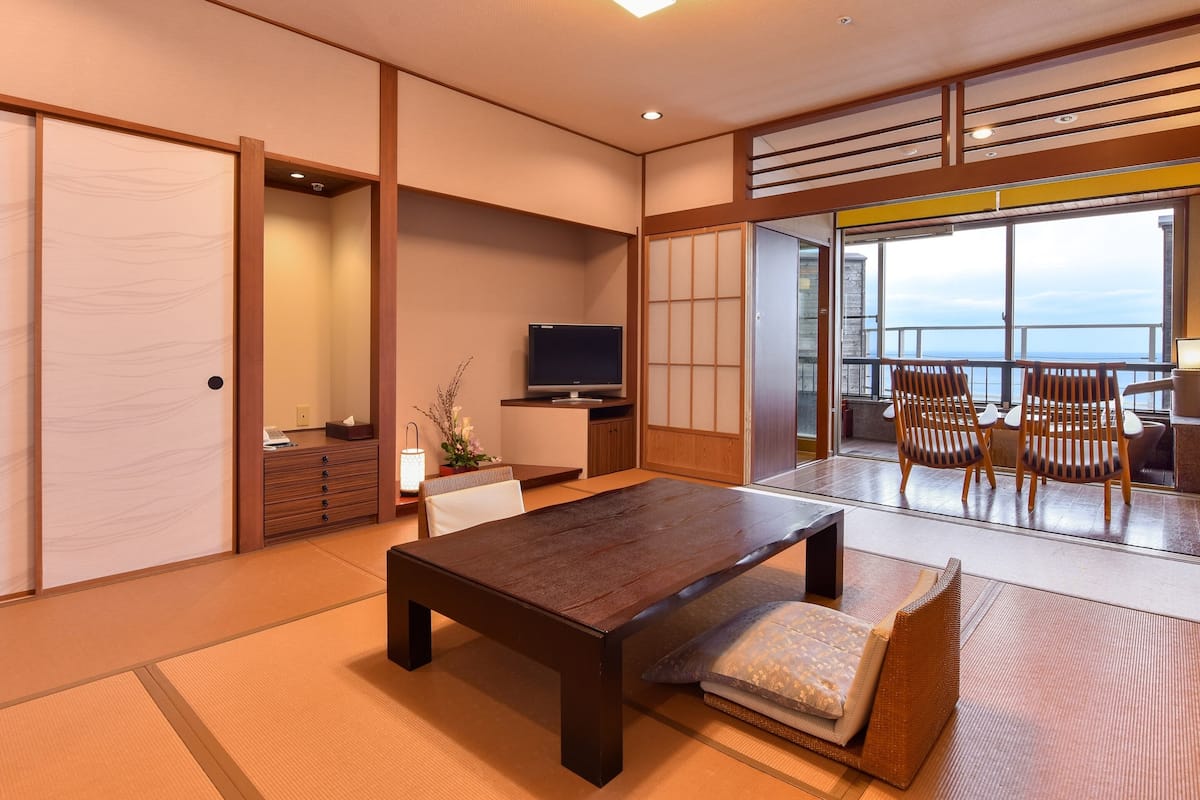 Una camera tradizionale in stile giapponese, con pavimento in tatami, un tavolino in legno, una televisione e una porta scorrevole che dà su un balcone con vista sul mare.