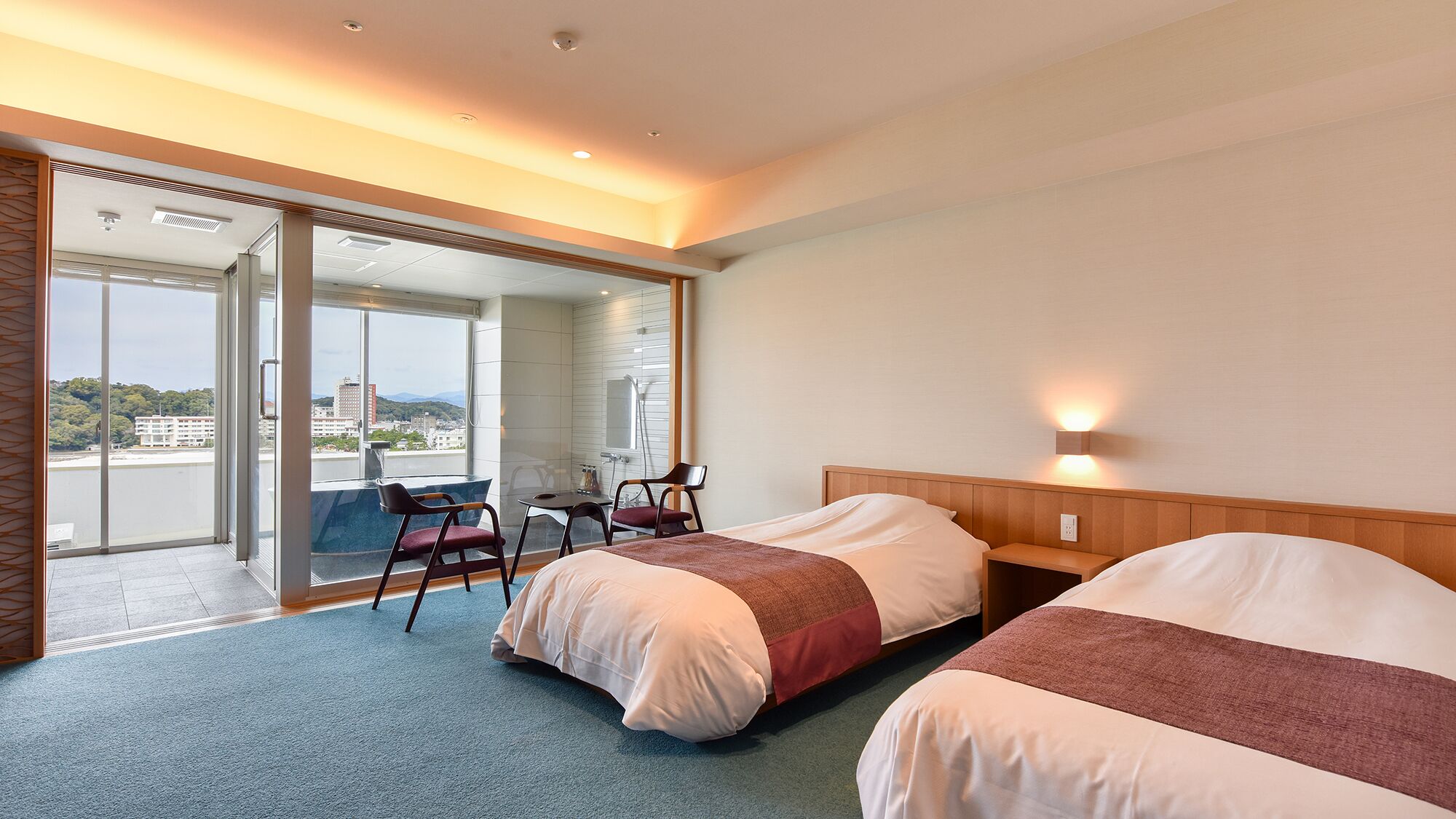 A hotel room with two beds, a desk, chairs, a large window with a view, and a sliding glass door.