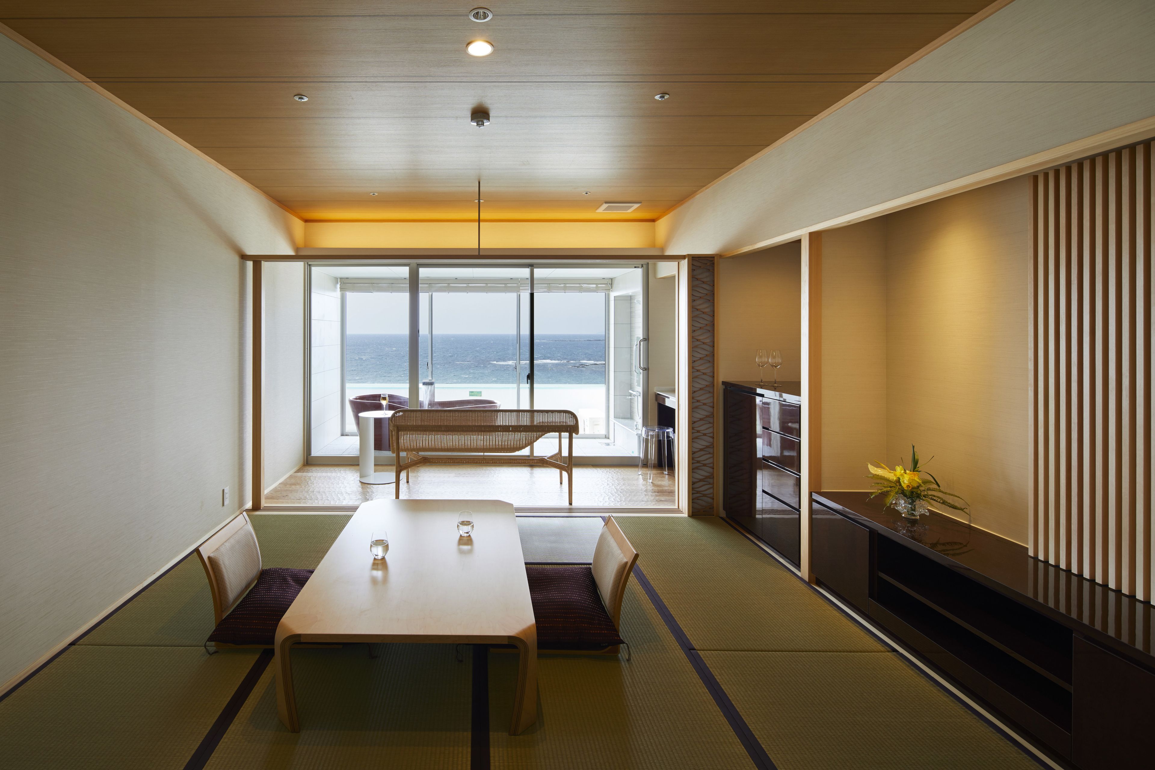 Une chambre moderne avec un plafond en bois, une grande porte-fenêtre coulissante donnant sur un balcon avec vue sur la mer, une table à manger avec des chaises et un téléviseur posé sur un meuble.