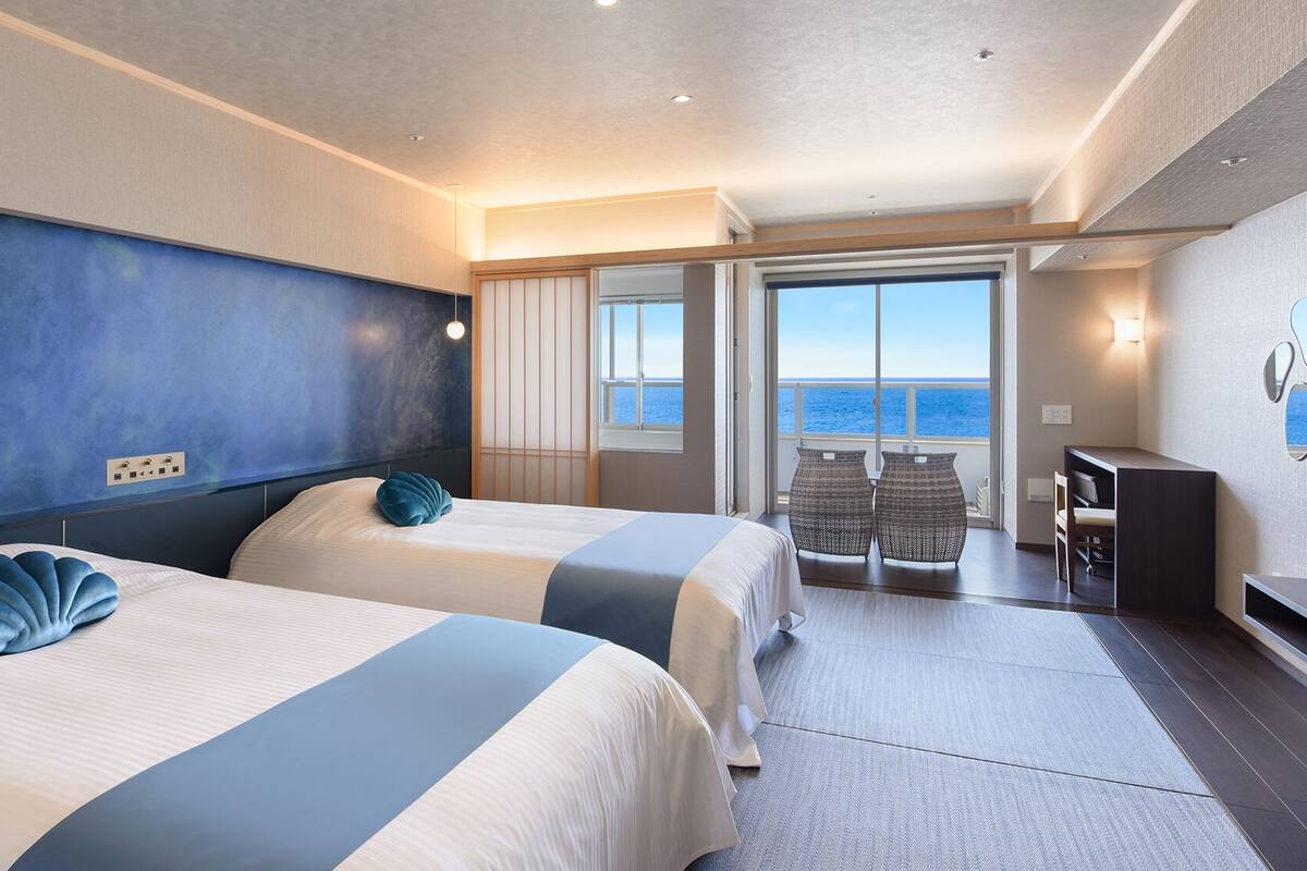 Camera d'albergo con due letti, vista mare, balcone con posti a sedere e una scrivania.