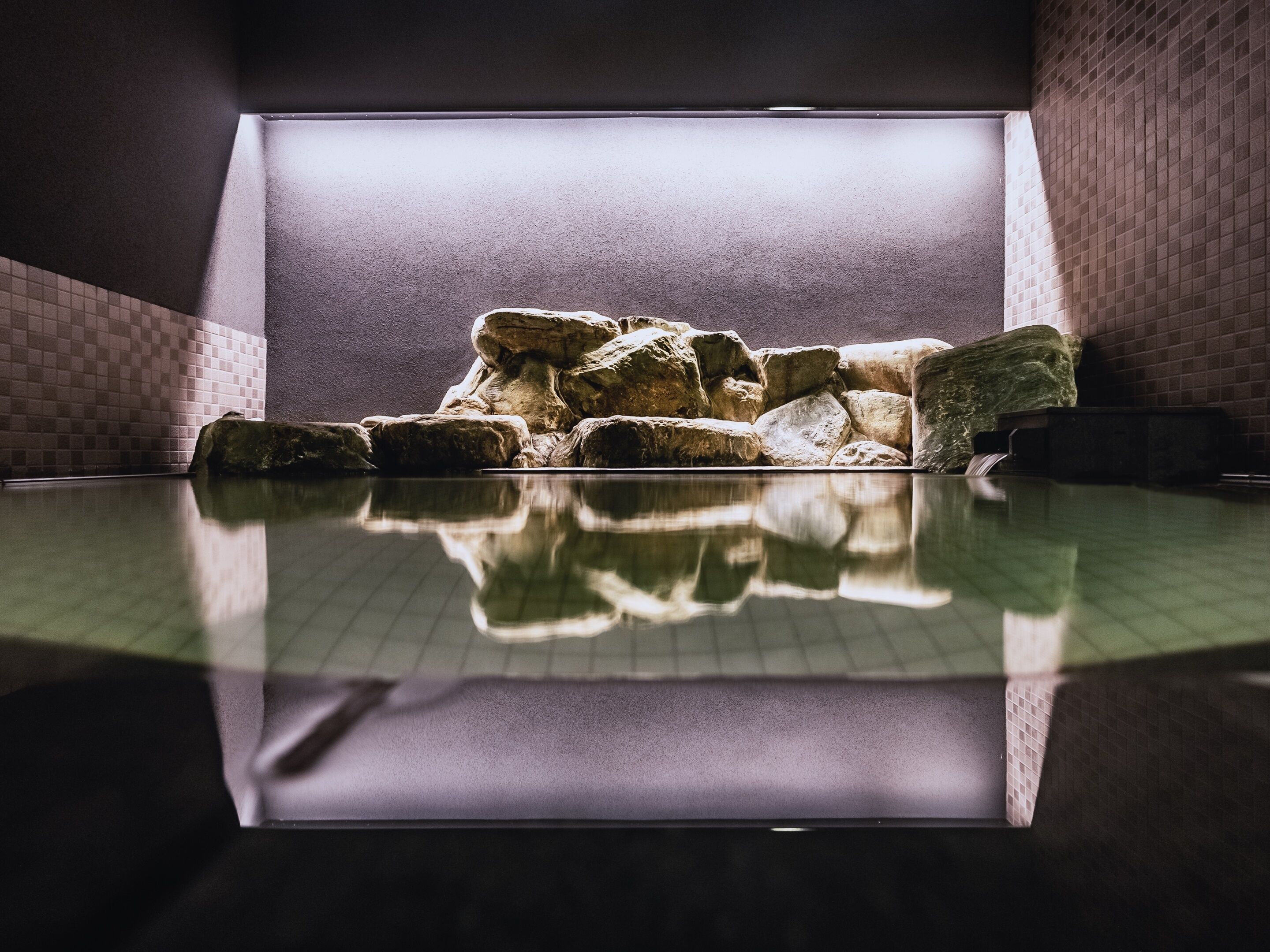 Une piscine dont l’eau est si claire qu’elle reflète l’environnement, entourée de murs en pierre et dotée d’un sol carrelé.