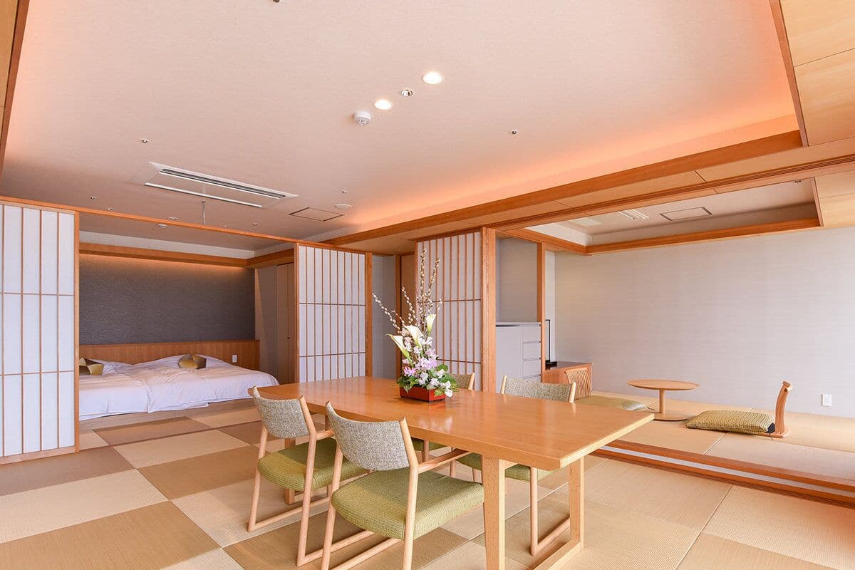 Una camera tradizionale in stile giapponese, con pavimento in tatami, un tavolino in legno con sedie, un letto con testiera in legno e un piccolo tavolino rotondo con un cuscino.