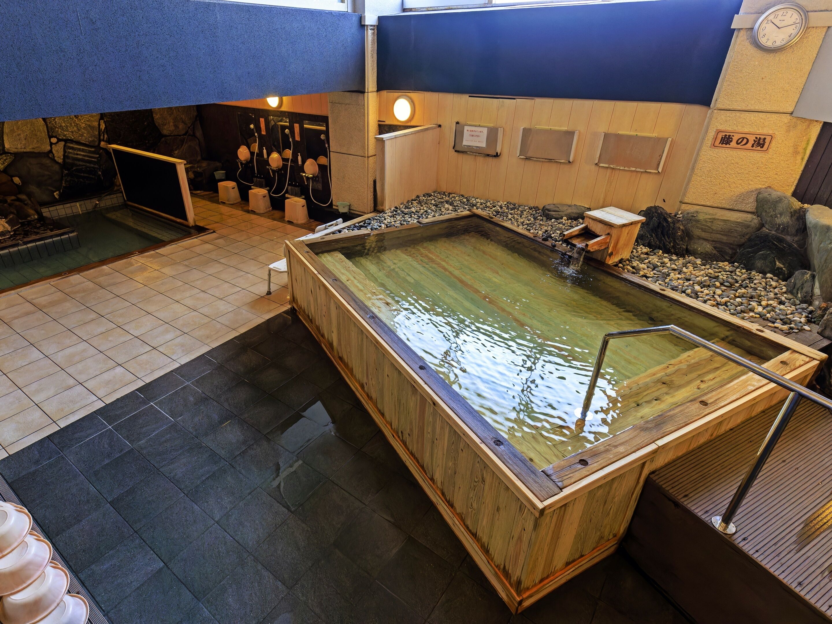 A traditional Japanese onsen with a wooden bath, stone seating, and a small waterfall.