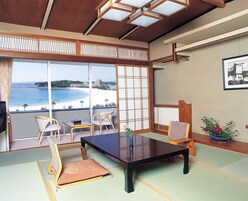 Una habitación japonesa tradicional con suelo de tatami, una mesa baja y sillas. Un ventanal ofrece vistas a una playa y edificios.