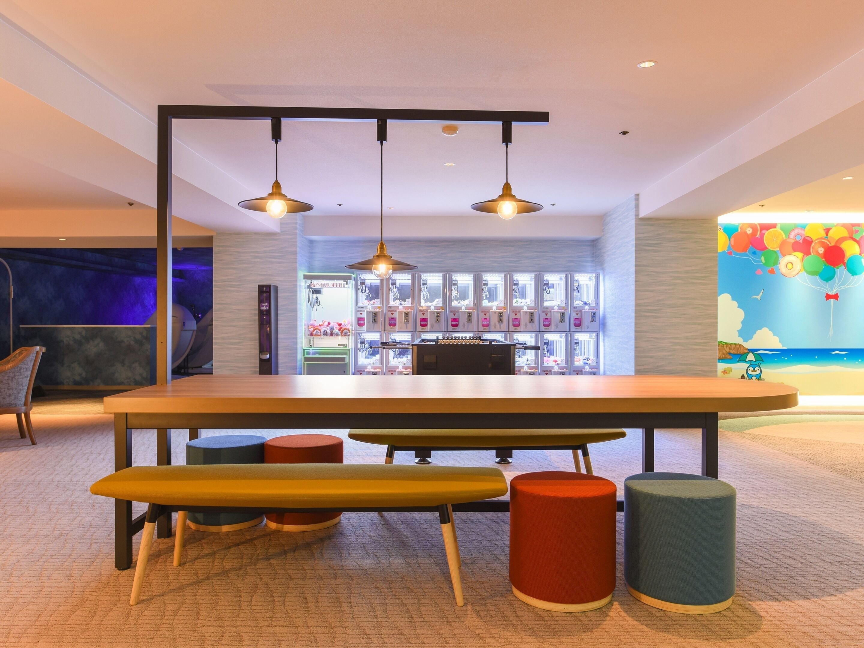 Um interior moderno com uma mesa comprida, banquetas coloridas e uma máquina de vendas automática ao fundo.