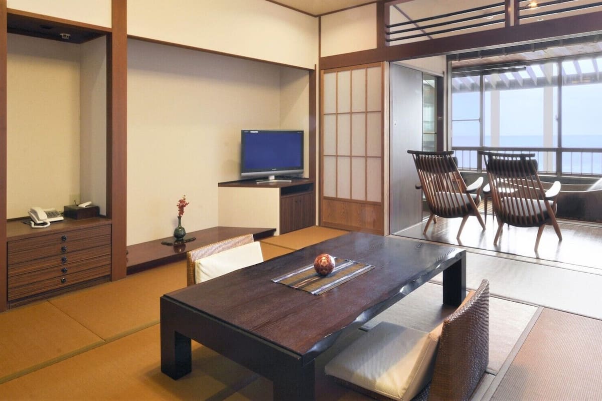 A traditional Japanese room with tatami flooring, a low table with cushions, a television, and a view of the sea.
