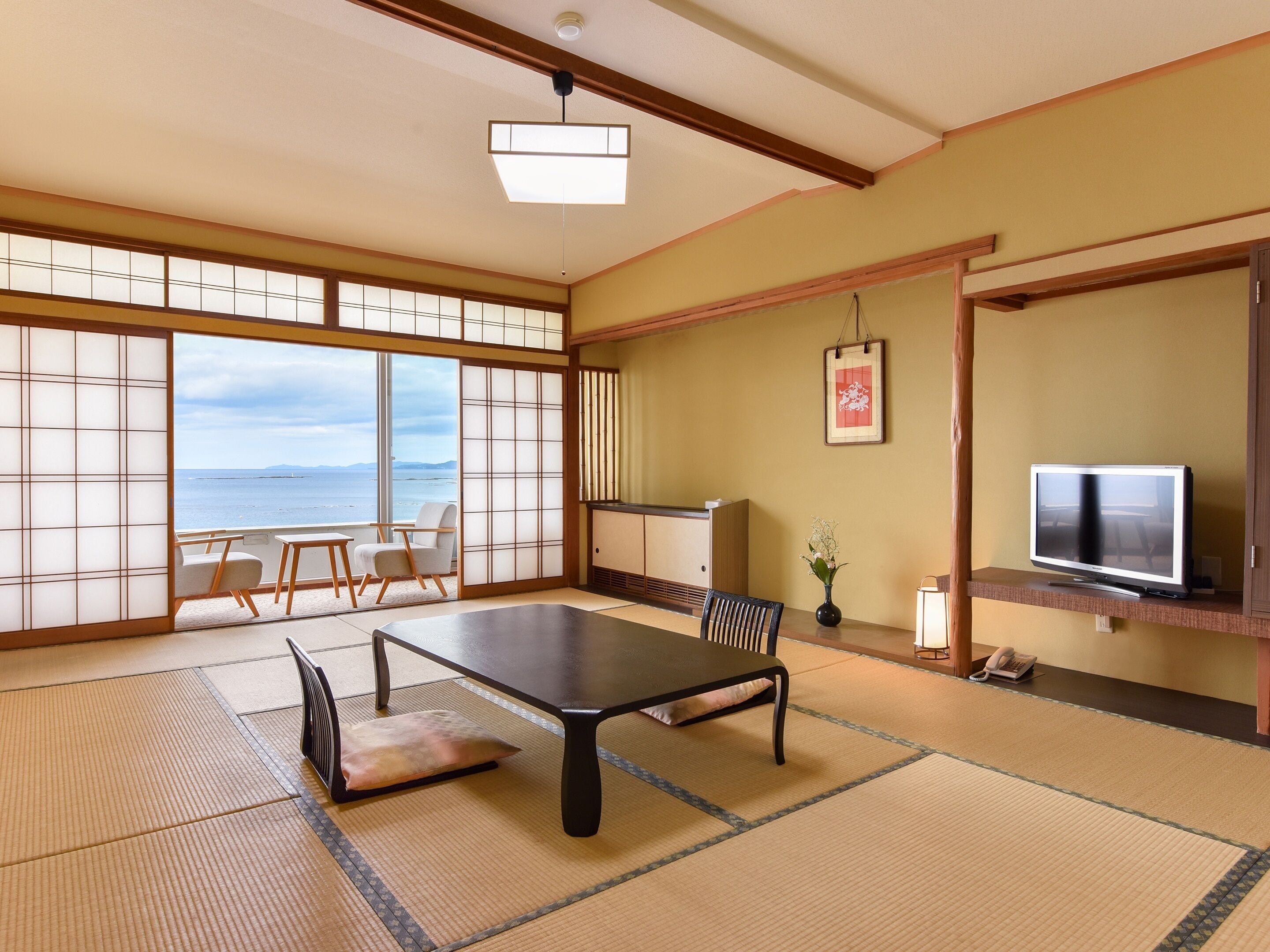 Una habitación japonesa tradicional con suelo de tatami, una mesa baja, sillas, un televisor y vista al mar.