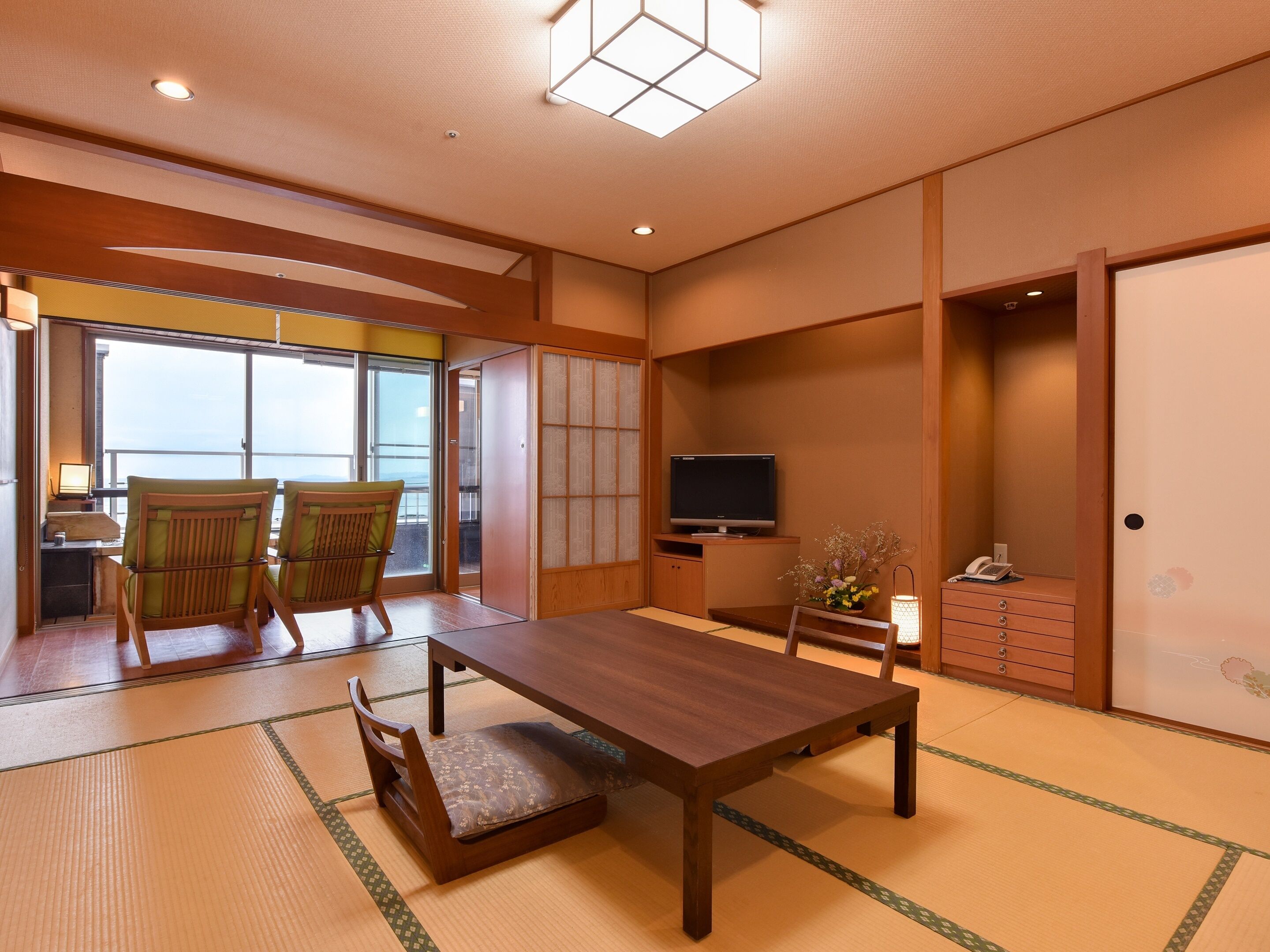 Una habitación tradicional japonesa con suelo de tatami, una mesa de madera baja, dos sillas con cojines verdes, un televisor y una puerta corrediza que da a un balcón.