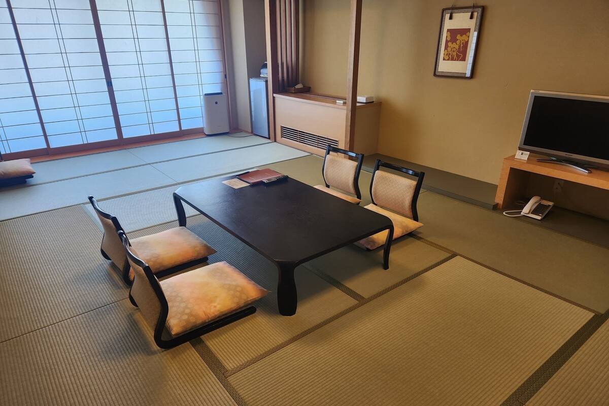 Una stanza con tatami, un tavolo nero, due sedie, un piccolo termosifone, una TV e un telefono su un supporto in legno.