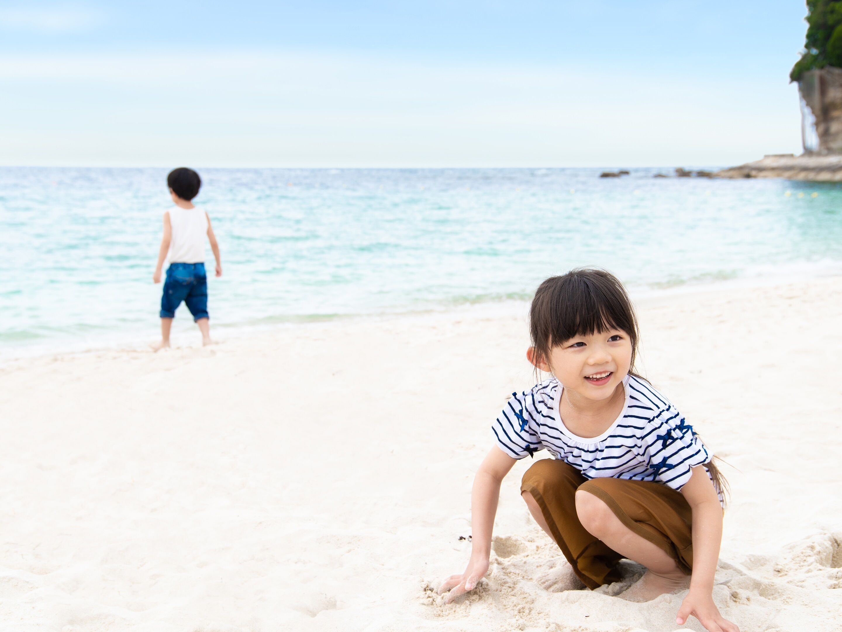 Escena de playa con un niño agachado en la arena y otro caminando al fondo.