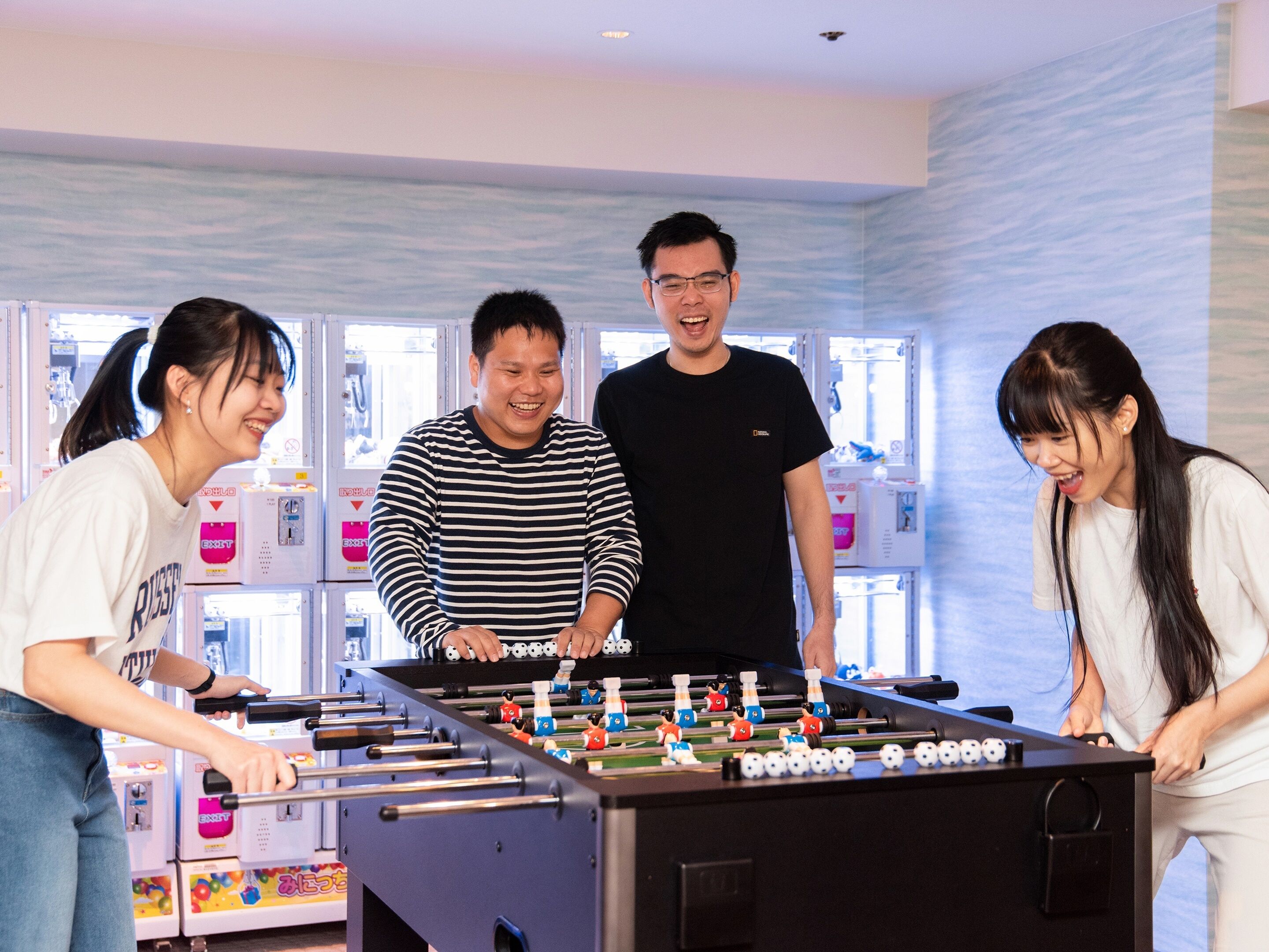 A group of people playing foosball in a room with a blue and white wall design and various gaming machines in the background.