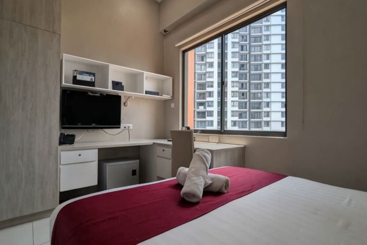 A bedroom with a bed, a desk, a chair, a television, and a window with a view of high-rise buildings.