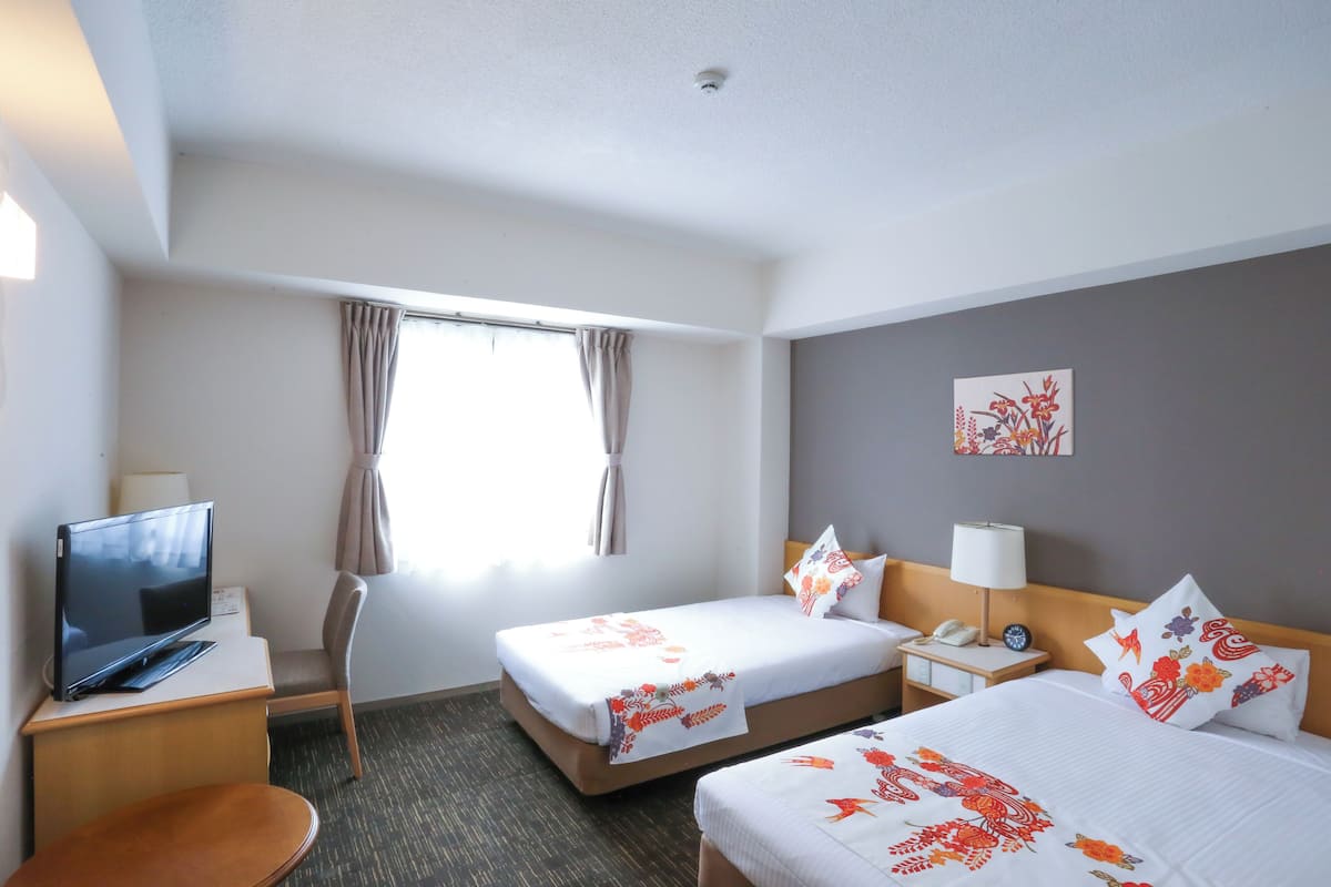 A hotel room with two beds, a desk with a TV, a chair, and a window with curtains.