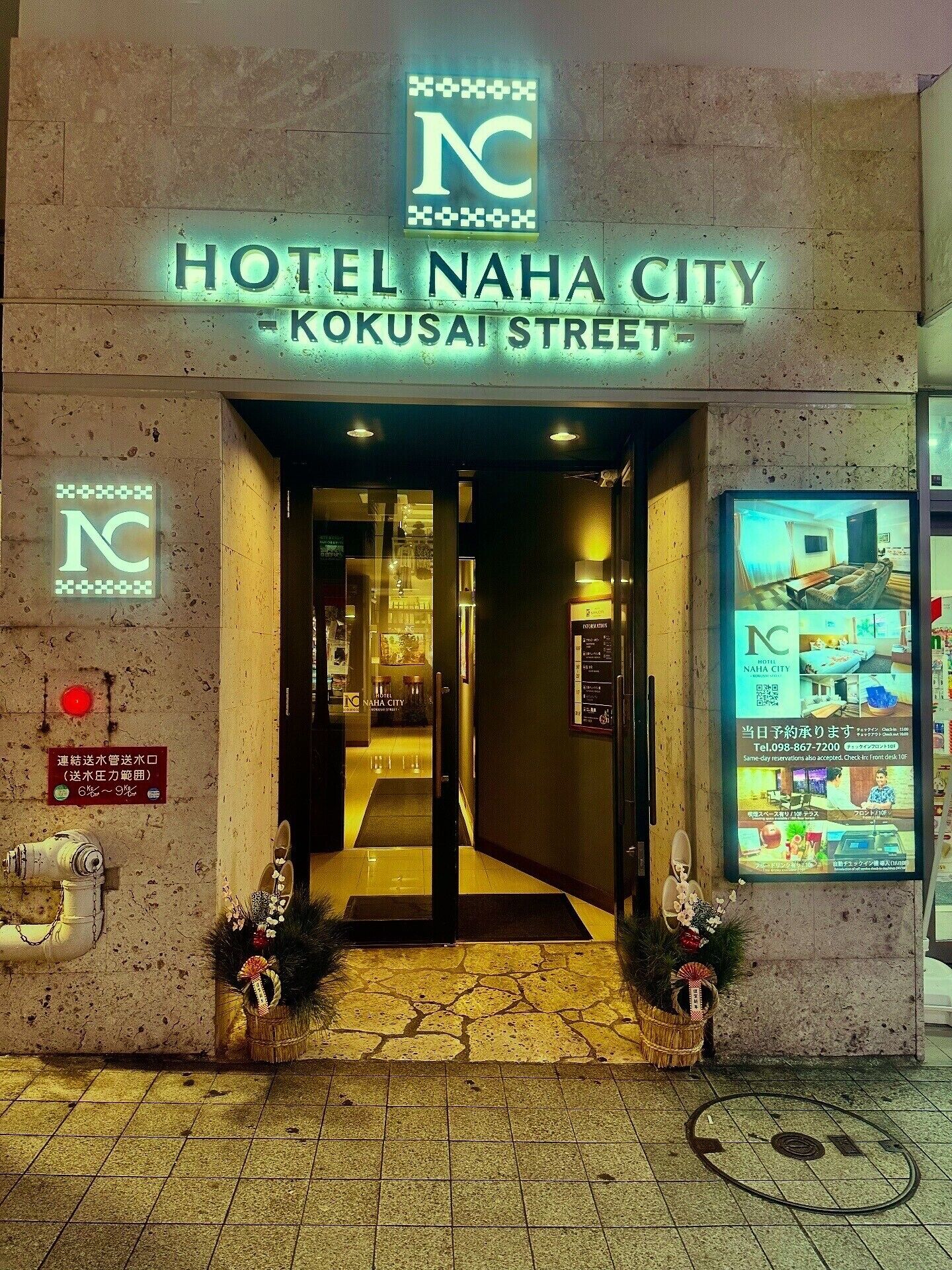 The entrance to HOTEL NAHA CITY on KOKUSAI STREET, featuring a neon sign, a reception desk, and a digital display with room images.