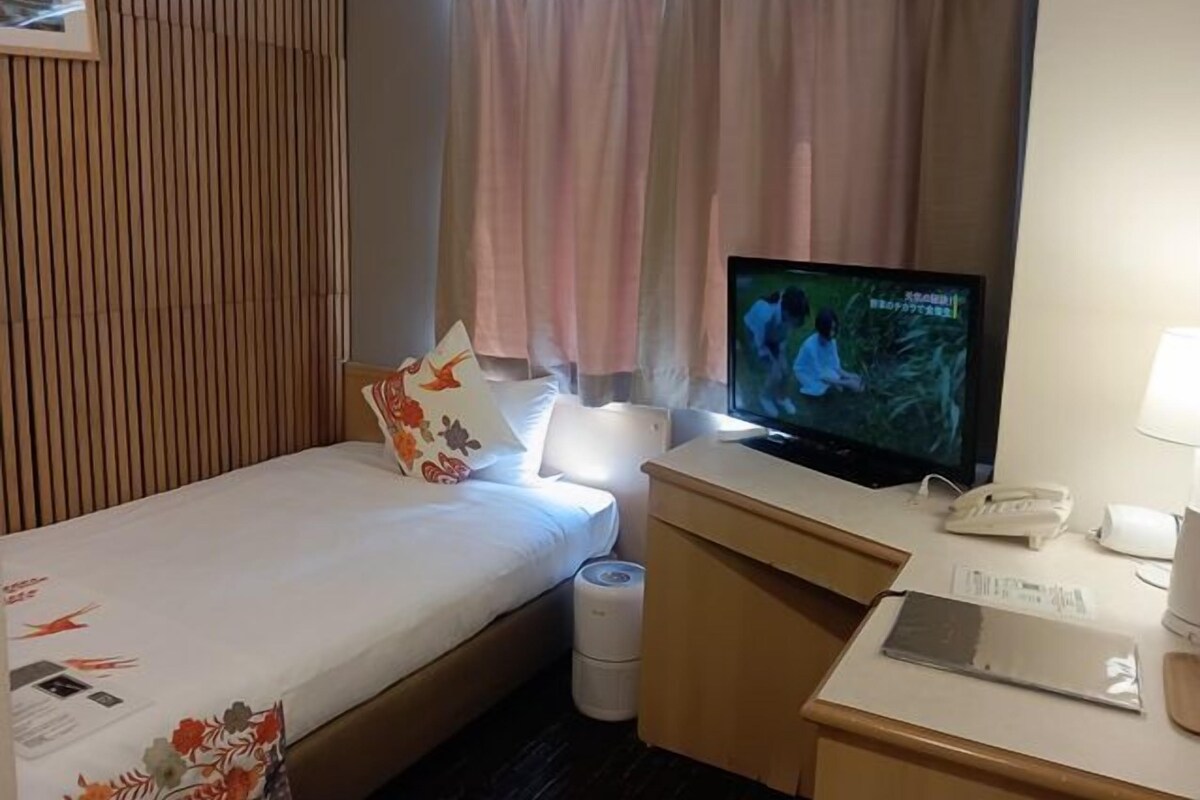 Ett hotellrum med en säng, ett skrivbord med en telefon, en tv och ett fönster med gardiner.