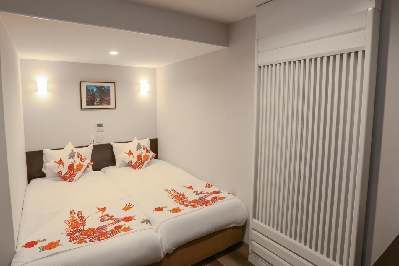A neatly made bed with white bedding and orange floral patterns, a framed picture on the wall, and a window with vertical blinds.