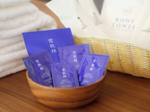 Una ciotola di legno contiene diversi sacchetti viola etichettati come 'Brightening Essence Lotion' e 'Foam Cleanser'. Sullo sfondo, un asciugamano bianco e un cesto con l'etichetta 'BODY TOWEL'.