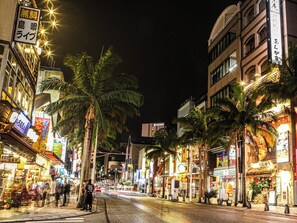 Scena notturna di una strada con palme, insegne illuminate e palazzi.