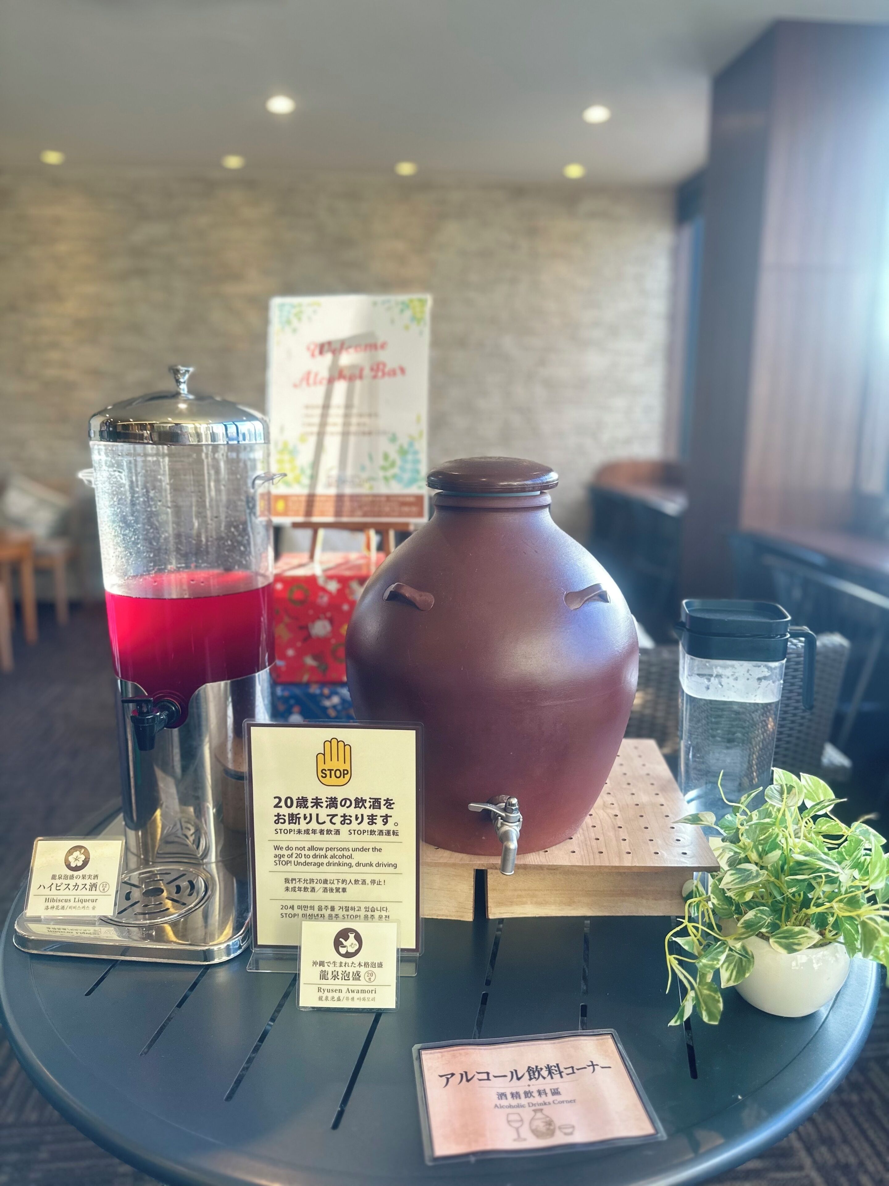 A table with a drink dispenser, a large ceramic pot, a water bottle, and a small potted plant. There are signs with text and a menu board in the background.