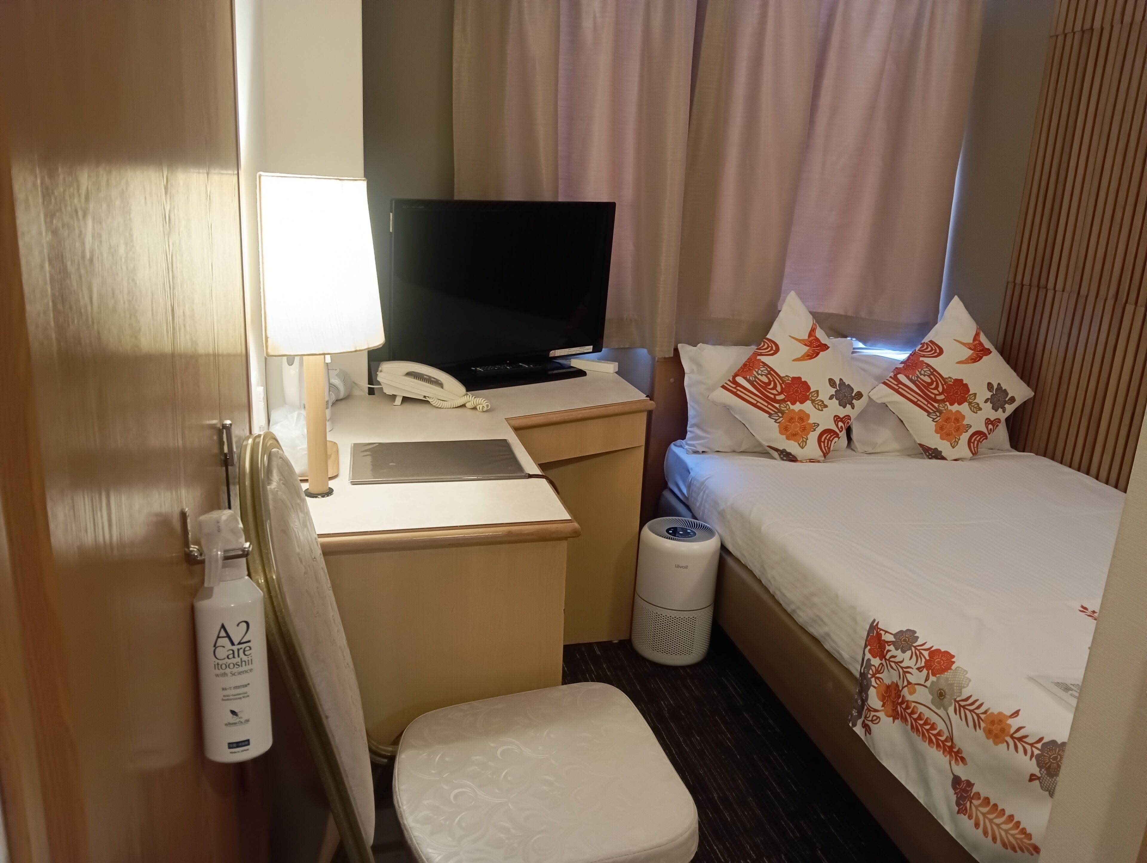 Een hotelkamer met een bed, een bureau met een computer, een telefoon, een lamp, een prullenbak en een fles A2 Care shampoo.
