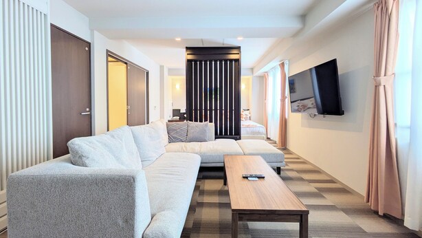A modern hotel room with a large sofa set, a wooden coffee table, a flat-screen TV mounted on the wall, and a view of the adjacent room. - Hotel Naha City -Kokusai Street- (Naha)