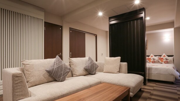 Une chambre d’hôtel moderne avec un canapé, une table basse en bois, un lit agrémenté d’oreillers à motifs floraux et un rideau foncé.