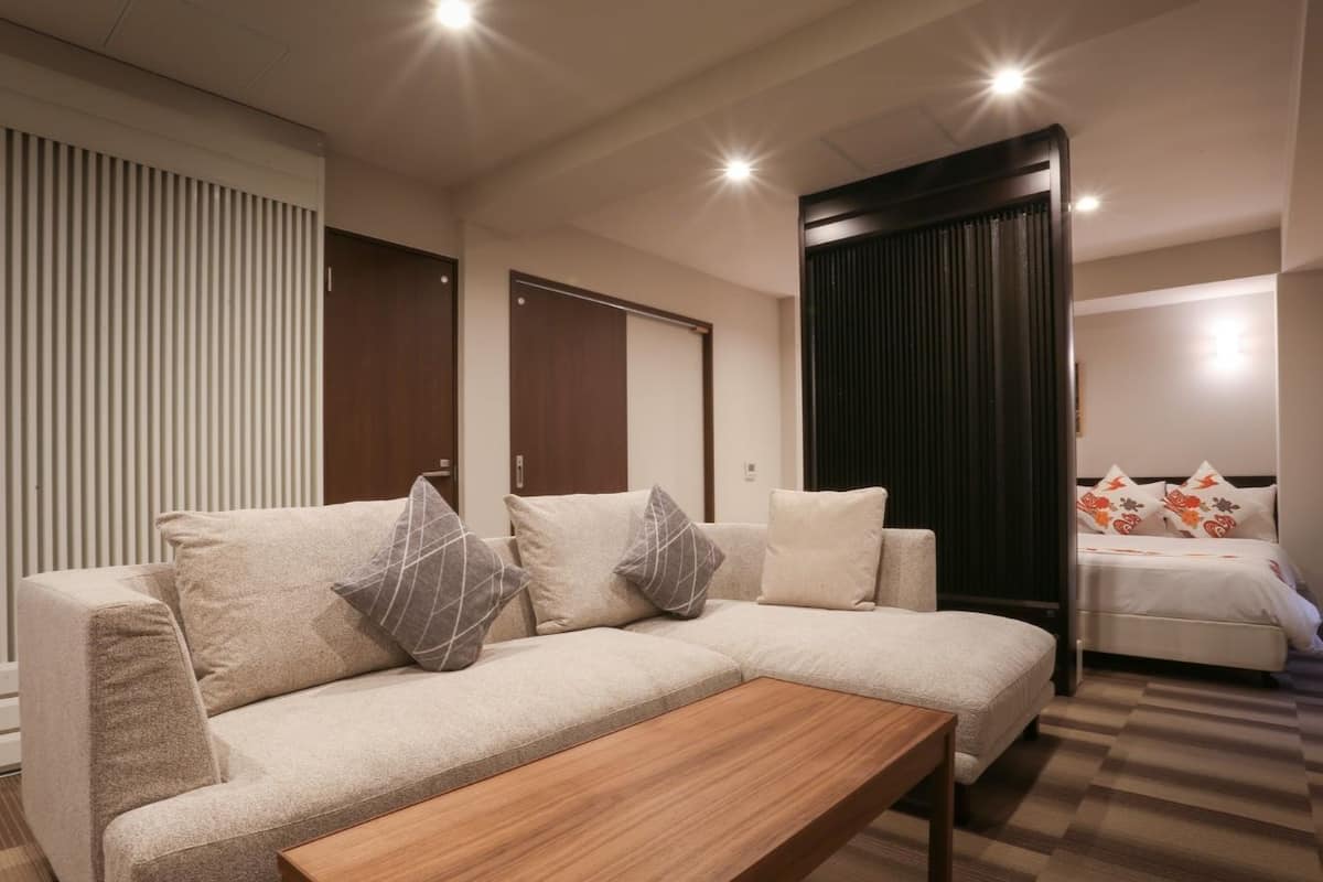A modern hotel room with a sofa, a wooden coffee table, a bed with floral pillows, and a dark curtain.