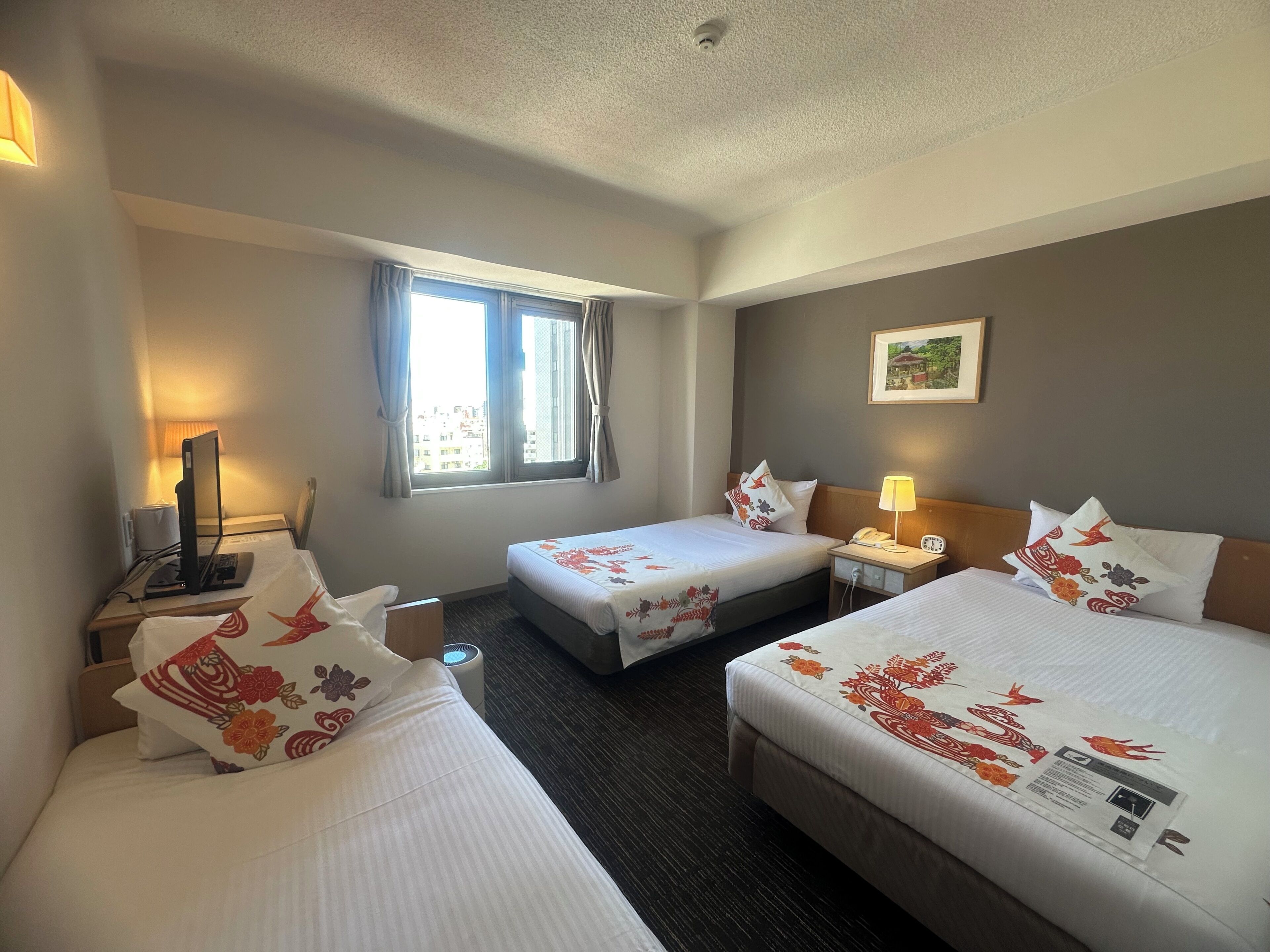 A hotel room with two beds, a desk with a computer, a window with curtains, a bedside lamp, and a wall-mounted picture.