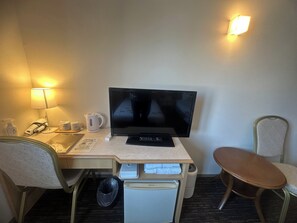 Habitación de hotel con escritorio, computadora, silla, refrigerador pequeño, cesto de basura y lámpara.