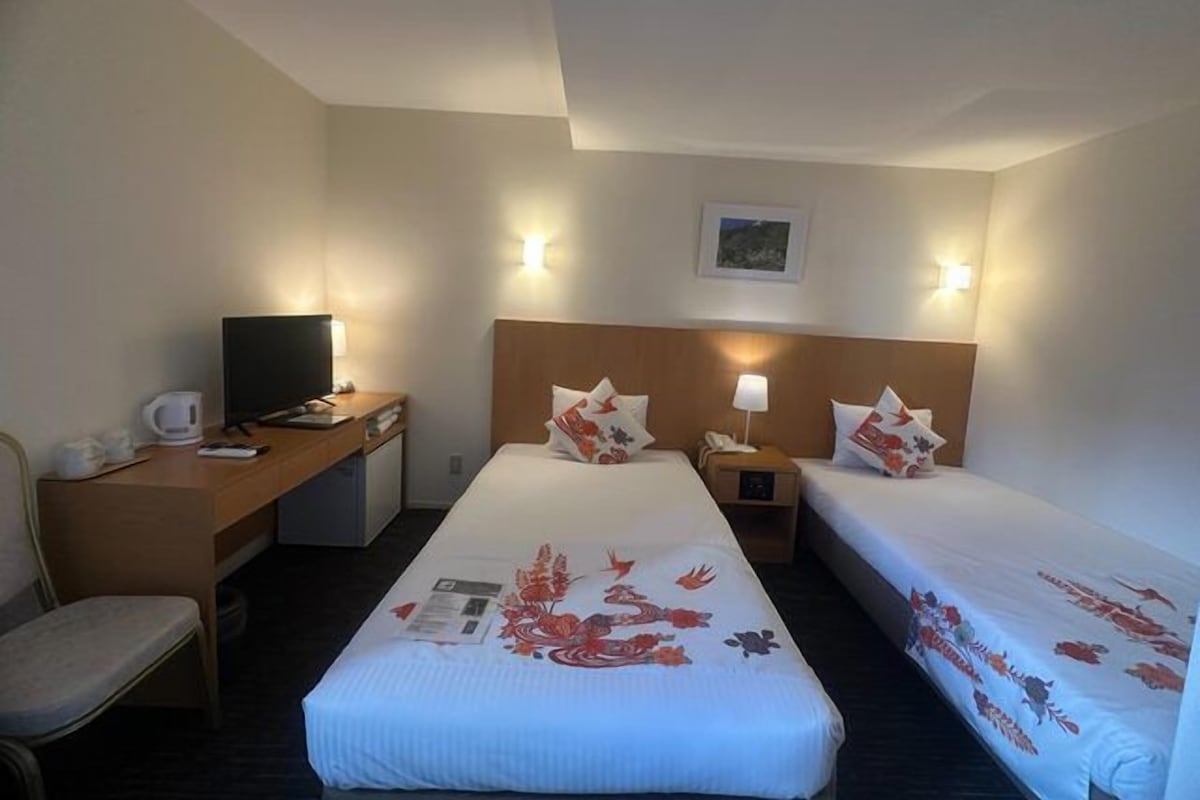 A hotel room with two beds, a desk with a computer, a chair, and a small table with a lamp.