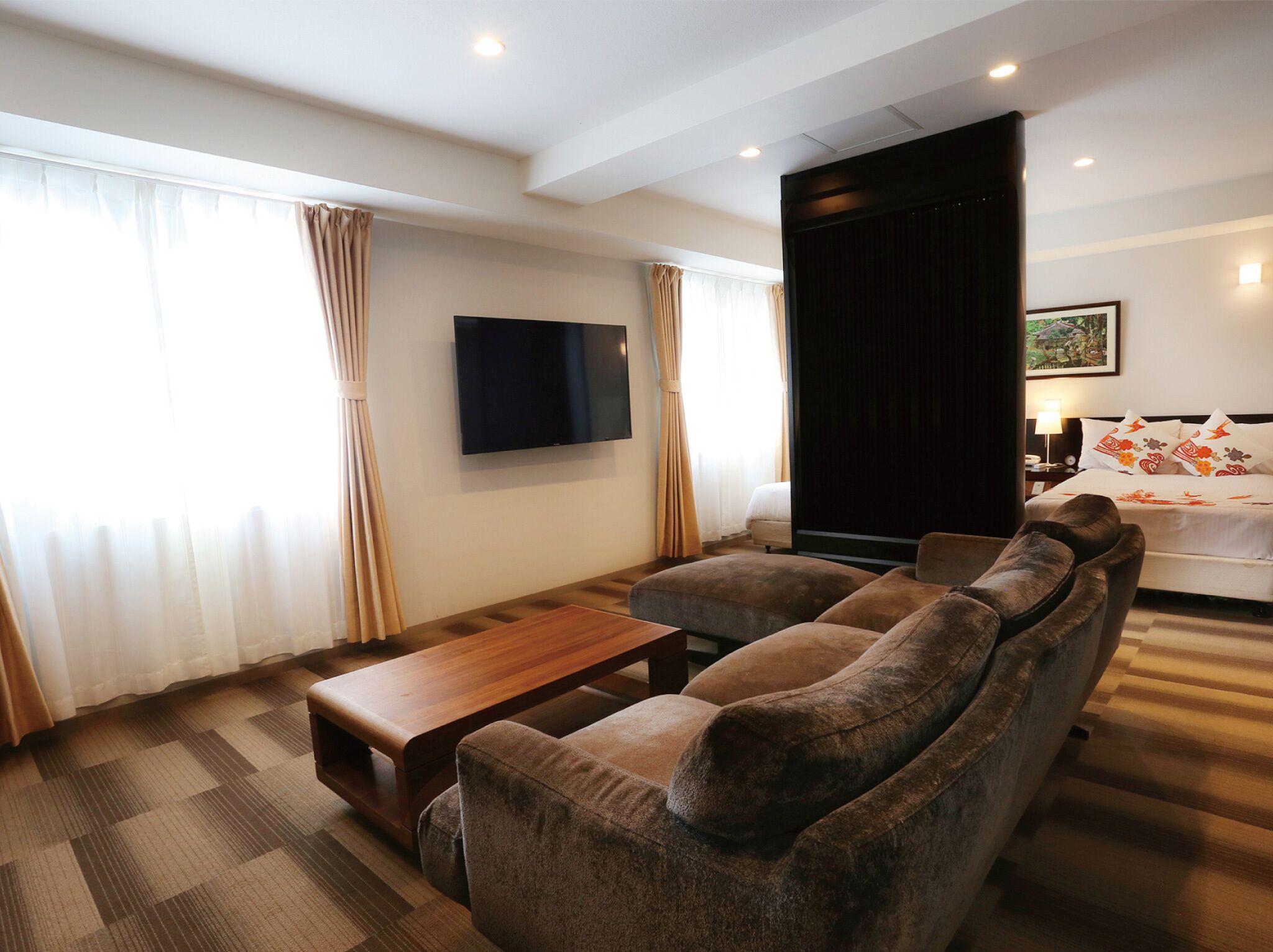 Quarto de hotel moderno com sofá, mesa de centro de madeira, TV de tela plana na parede e cama com almofadas decorativas.