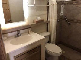 Un baño con lavamanos blanco, espejo e inodoro. Hay una cortina de ducha y una barra de apoyo en la zona de la ducha.