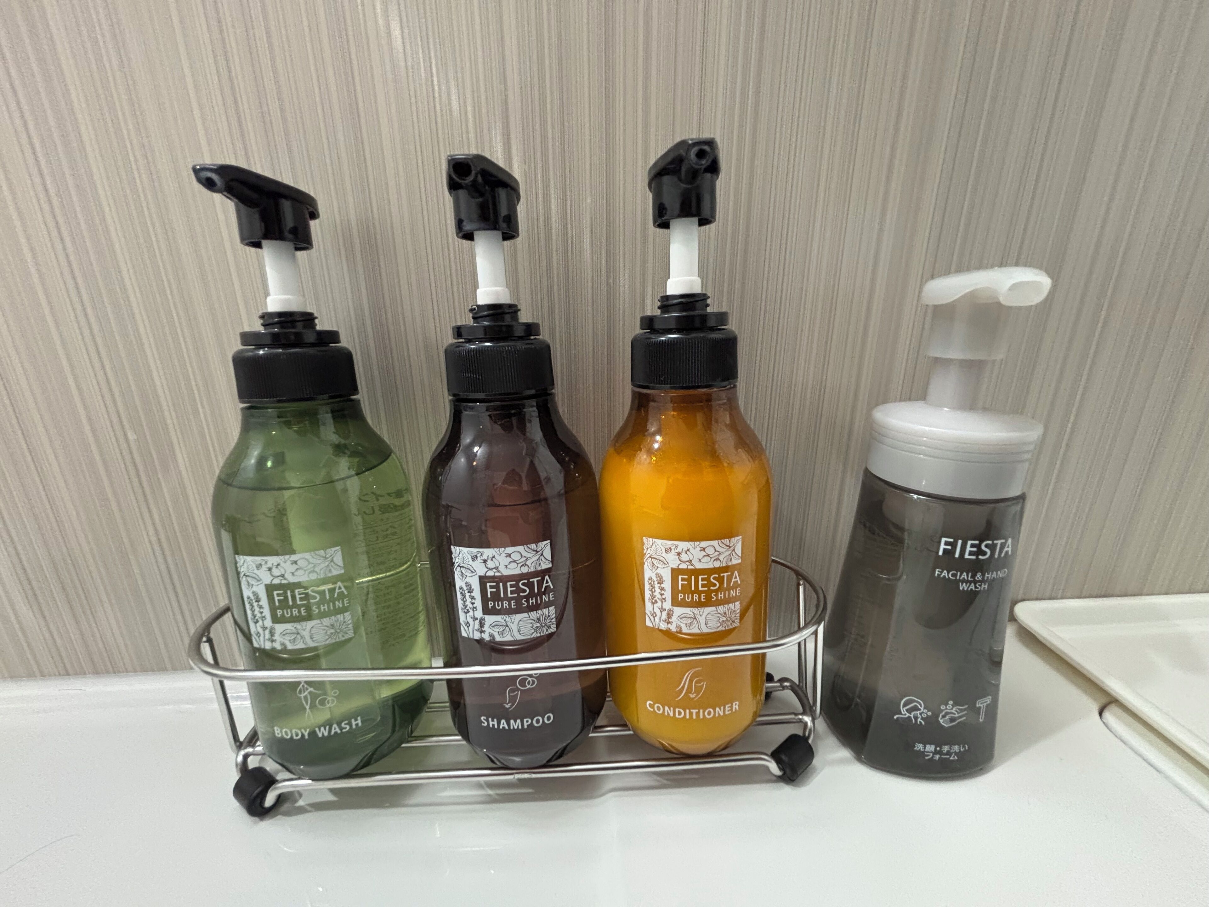 Una estantería de metal con cuatro botellas de productos de cuidado personal Fiesta: jabón corporal, champú, acondicionador y limpiador facial y de manos.