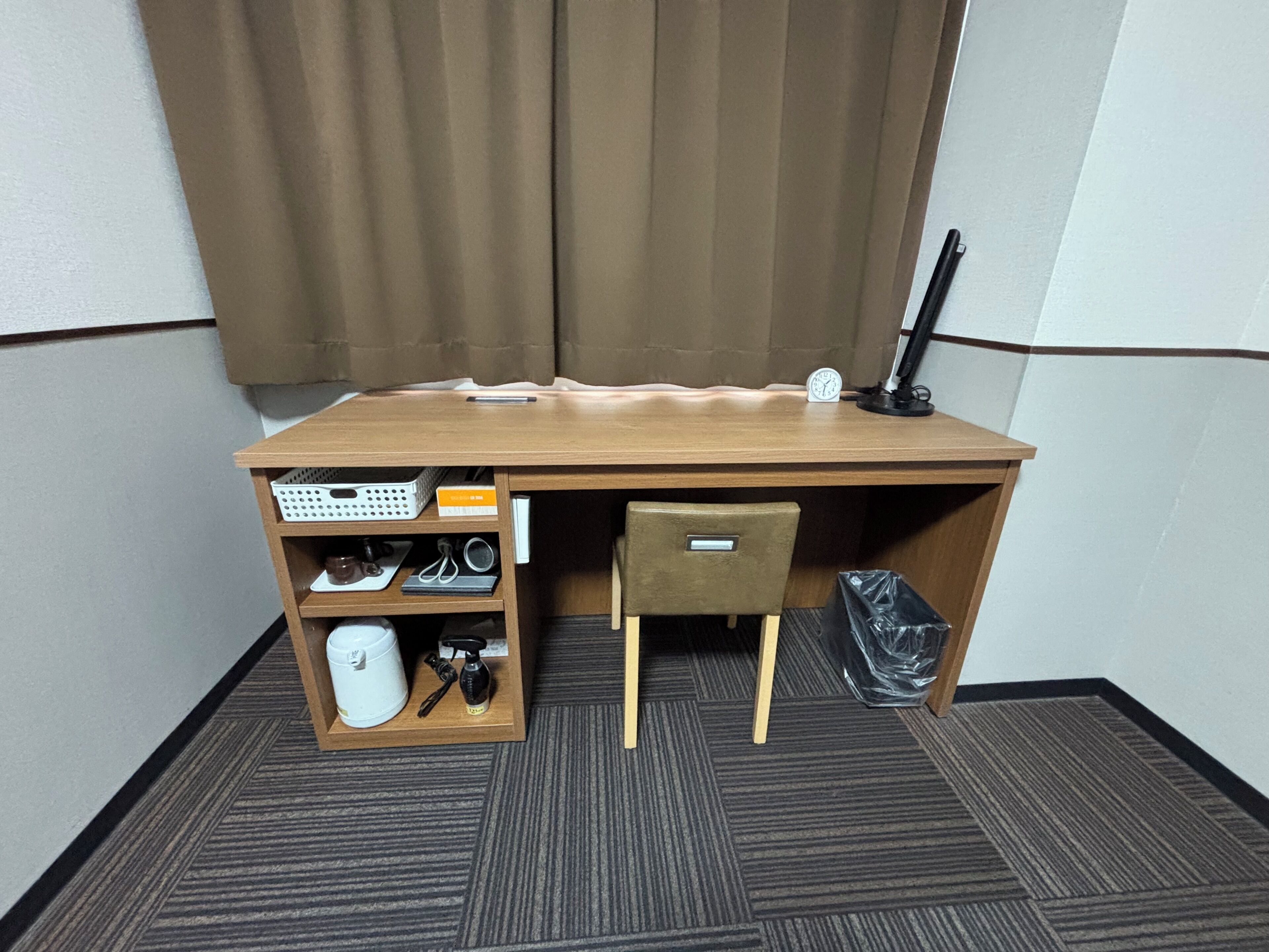 Un espacio de oficina pequeño con un escritorio de madera, una silla, un estante con varios objetos, un cubo de basura y un reloj en la pared.