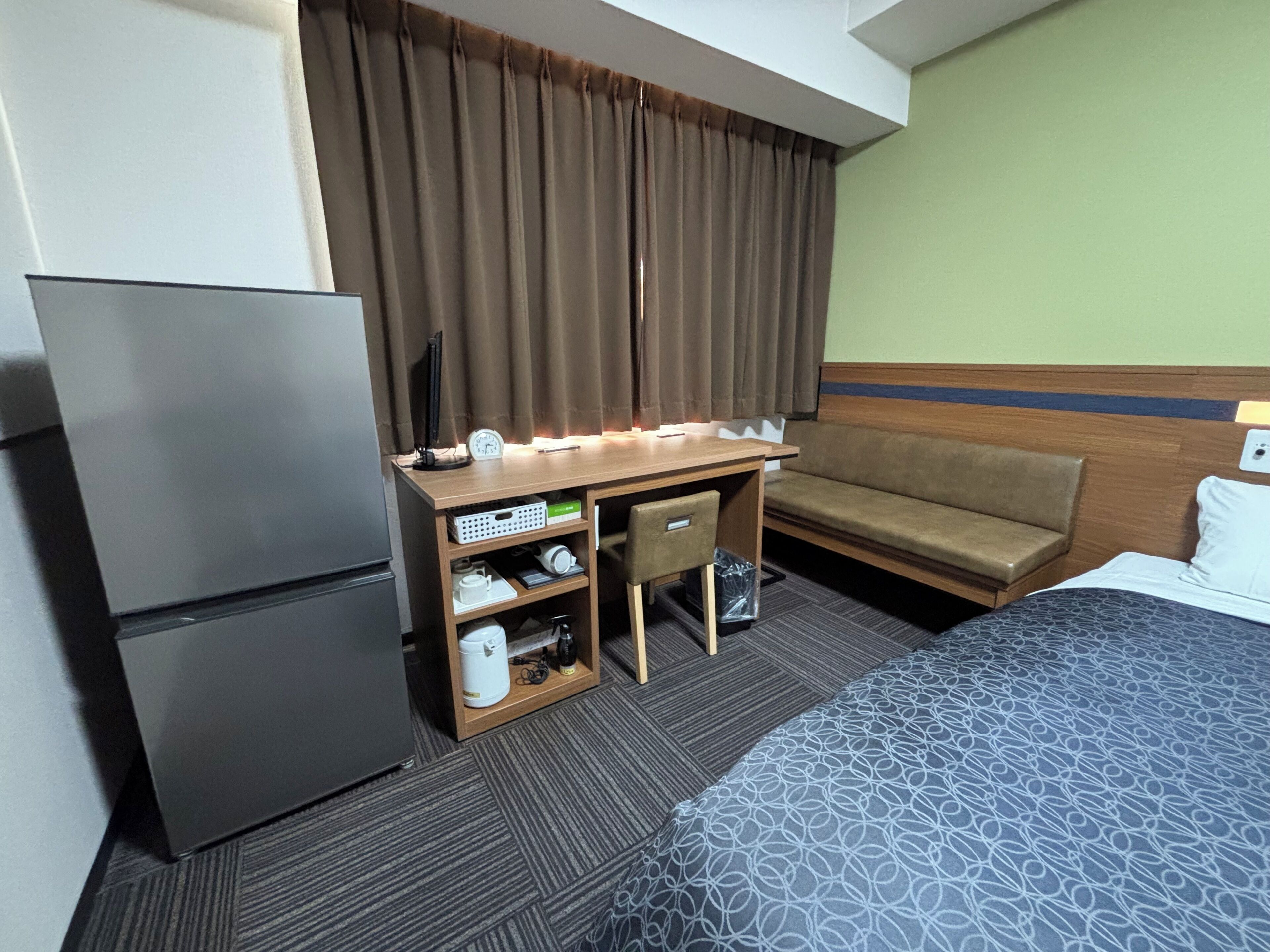 A hotel room with a bed, a desk with a computer, a refrigerator, a sofa, and a window with curtains.