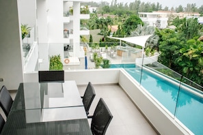 Uma área moderna para refeições ao ar livre, com mesa de vidro e cadeiras de vime, com vista para uma piscina e vegetação exuberante.