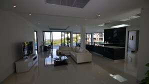 A modern living room with a sofa, coffee table, and TV. There is a kitchen area with a large island and a dining area with chairs. The room has large windows and a view of the outdoors. - Sansuri Resort Phuket (Choeng Thale)