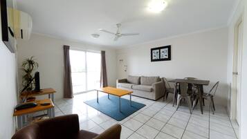 A living room with a sofa, a coffee table, a dining table with chairs, a ceiling fan, and a large window with curtains.