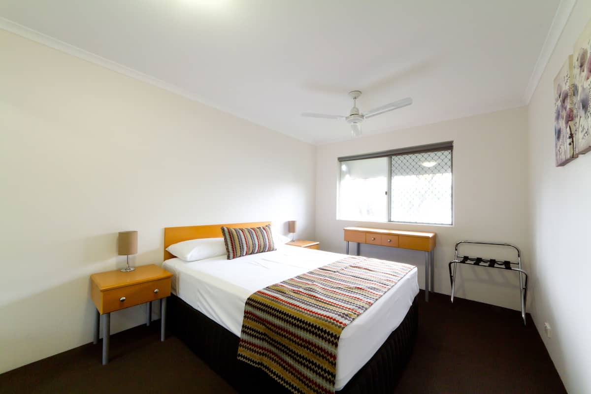 A hotel room with a bed, a nightstand, a desk, a window, and a ceiling fan.