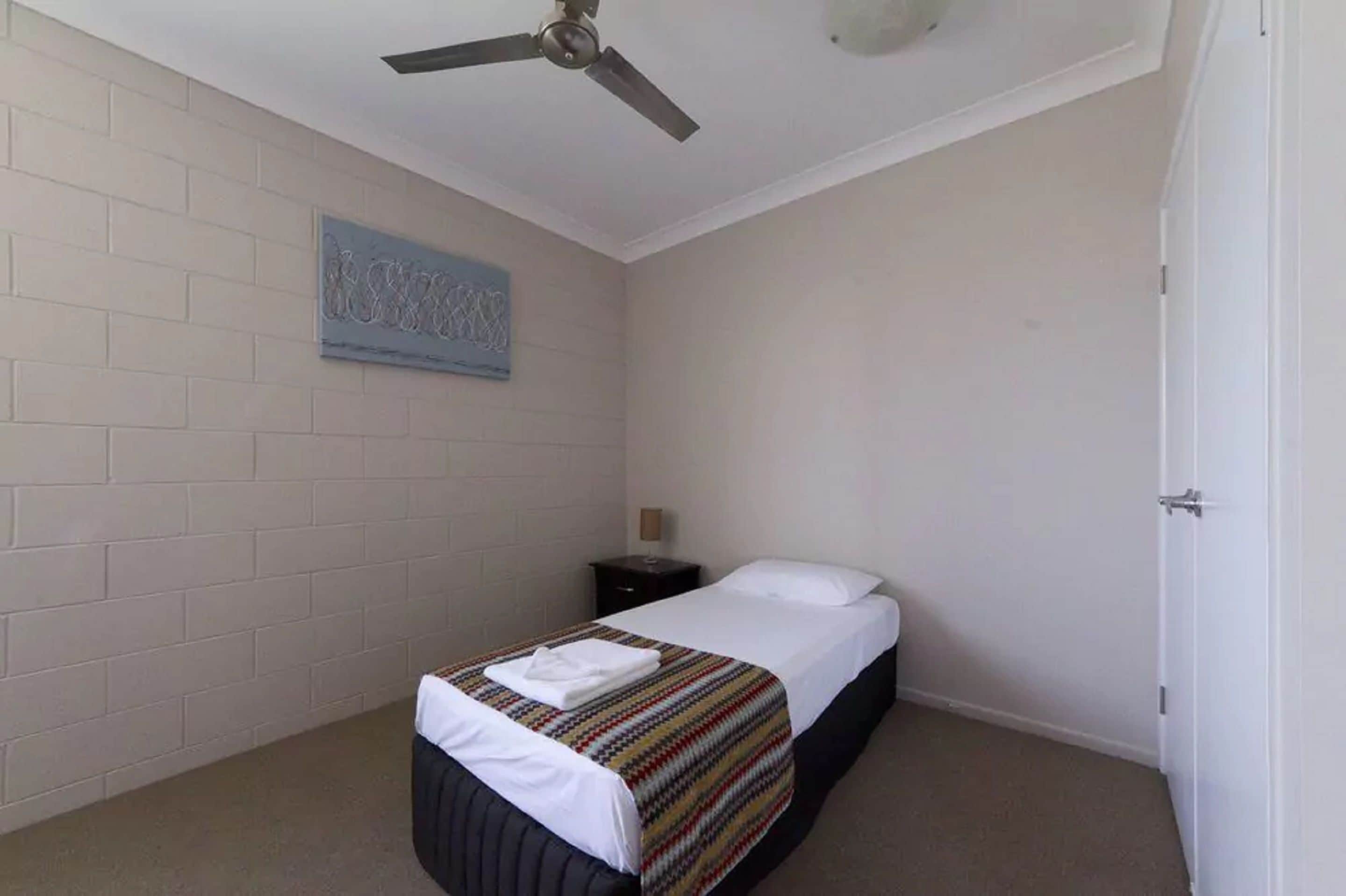 Uma cama de solteiro com um cobertor listrado e travesseiro branco, uma pequena mesa de cabeceira, um ventilador de teto e um interruptor de luz na parede.