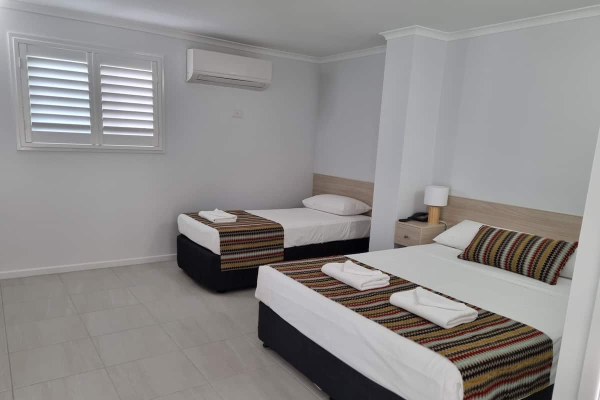 A hotel room with two beds, a window with blinds, an air conditioning unit, and a bedside table with a lamp.