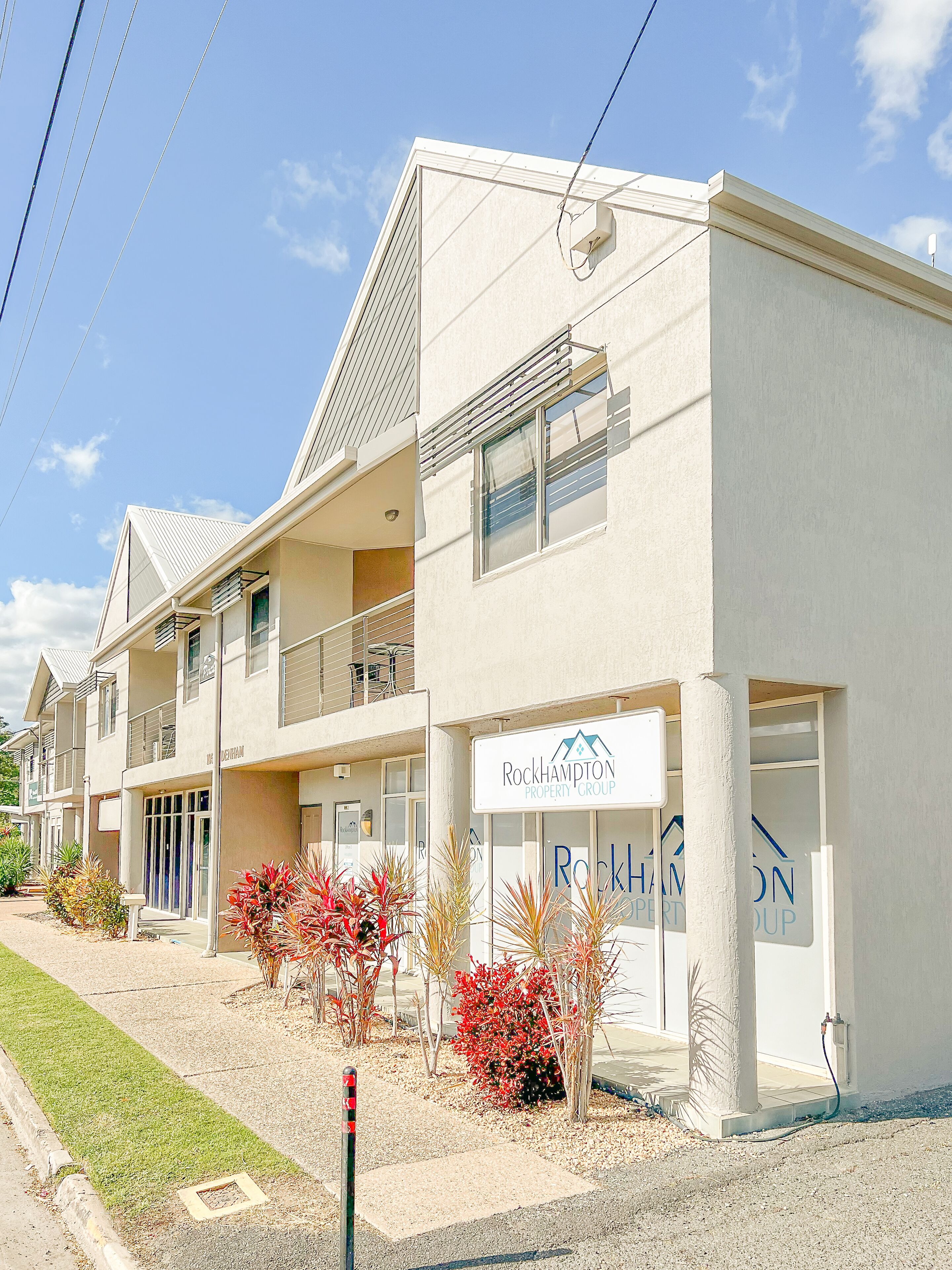 A multi-story building with a sign for Rockhampton Property Group. It features balconies, a gated entrance, and a landscaped front yard with shrubs.