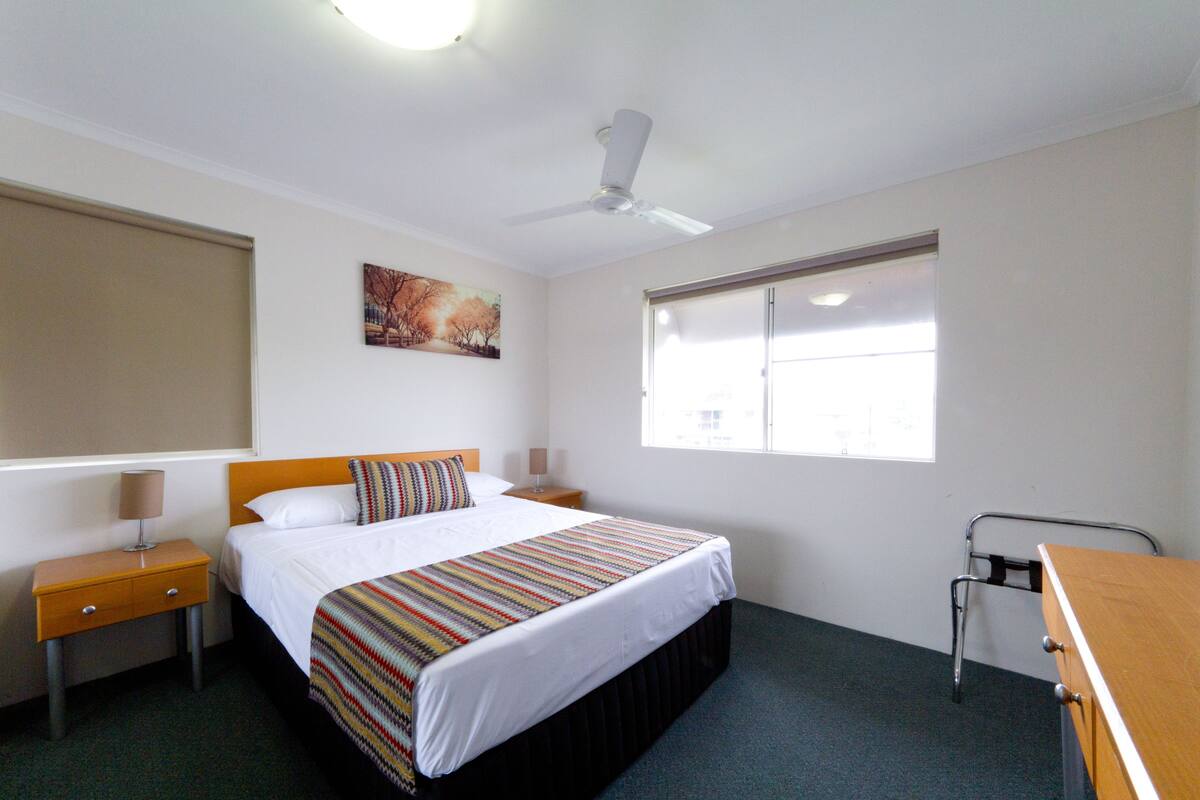 A hotel room with a bed, a bedside table, a lamp, a window with blinds, a ceiling fan, and a painting on the wall.